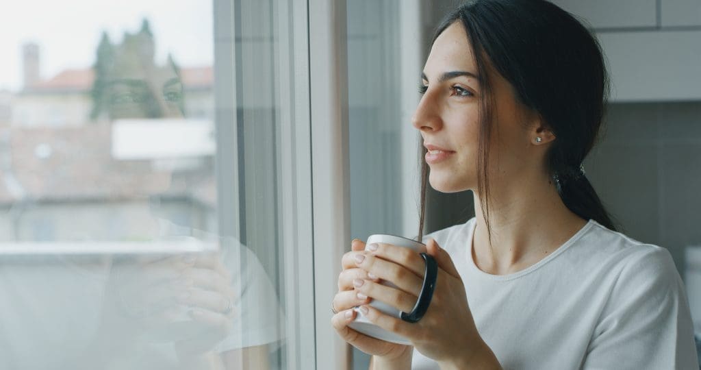 Woman holding a mug of coffee looking out the window smiling