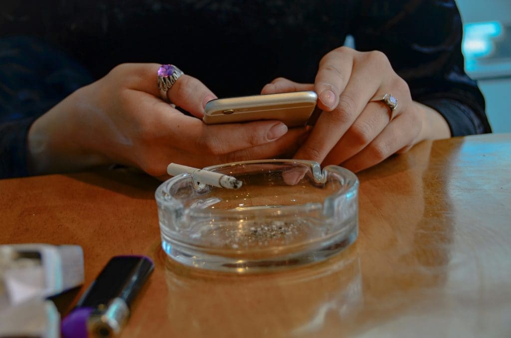 cigarette in a clear ash tray and someone gambling on their phone